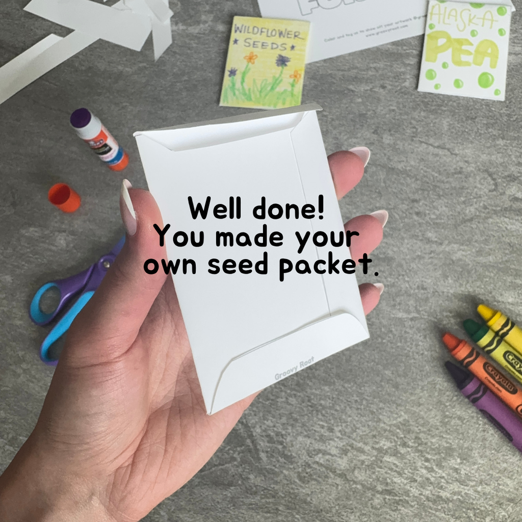 Hand holding a white seed packet with text 'Well done! You made your own seed packet.' on a gray surface with craft supplies.