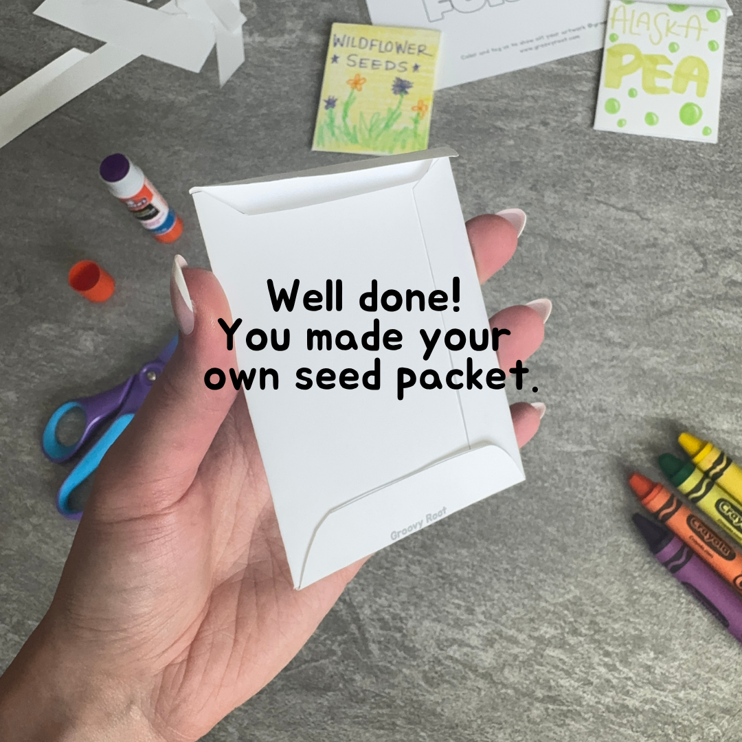 Hand holding a white seed packet with text 'Well done! You made your own seed packet.' on a gray surface with craft supplies.