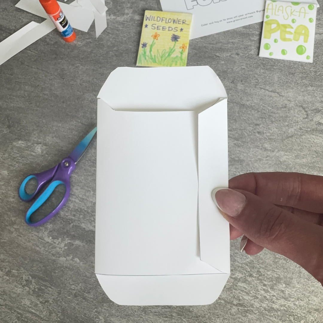 Blank seed packet held by a hand on a gray surface with scissors and seed packets in the background.