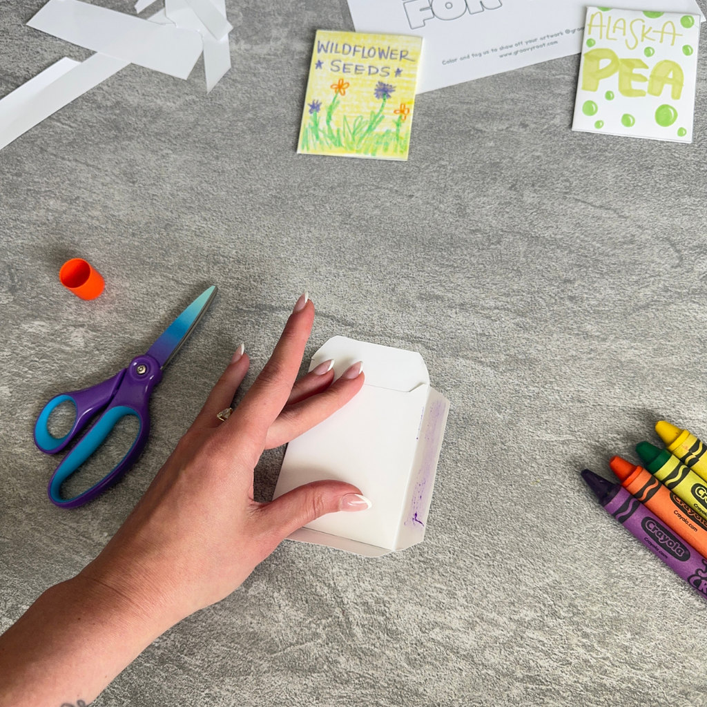 Hand holding a white paper craft on a gray surface with scissors, crayons, and seed packets.