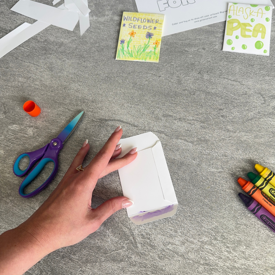 Hand holding a blank seed packet on a gray surface with scissors, crayons, and seed packets in the background.