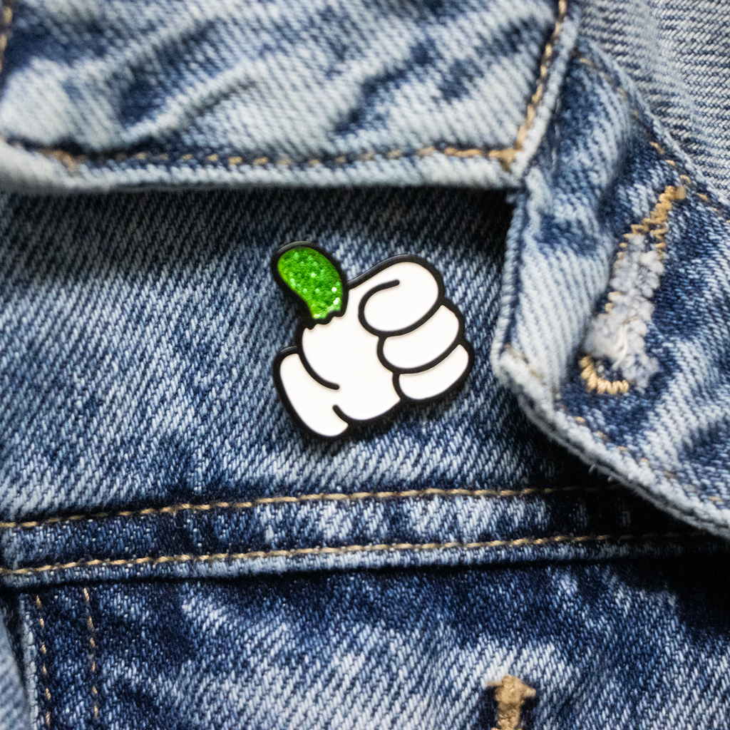 An enamel pin in the shape of a "thumbs up" with a sparkling green thumb on a denim jacket.