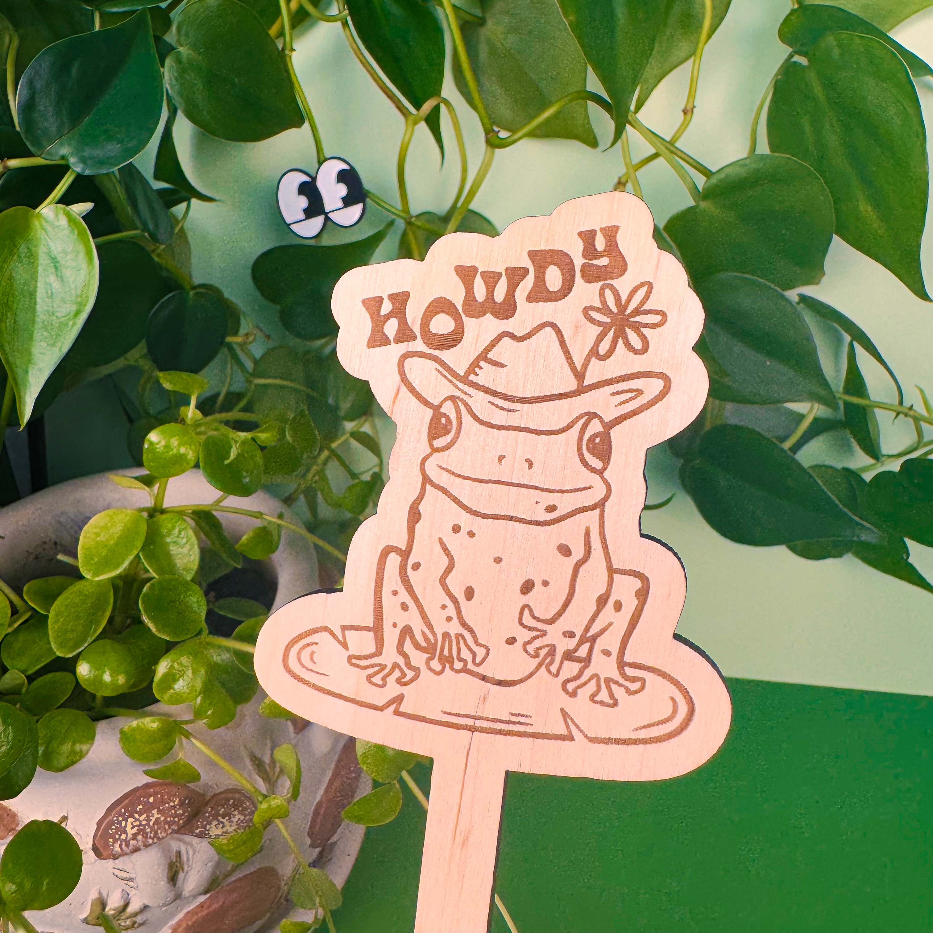 Decorative plant stake with a frog design and 'Howdy' text, surrounded by green leaves.