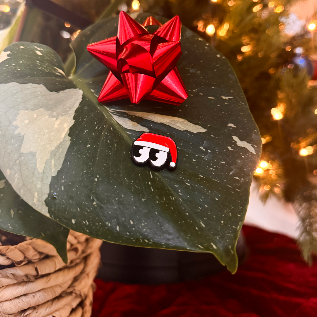 Red star-shaped bow on a large green leaf with a Christmas-themed face emoji in the foreground, against a blurred festive background.
