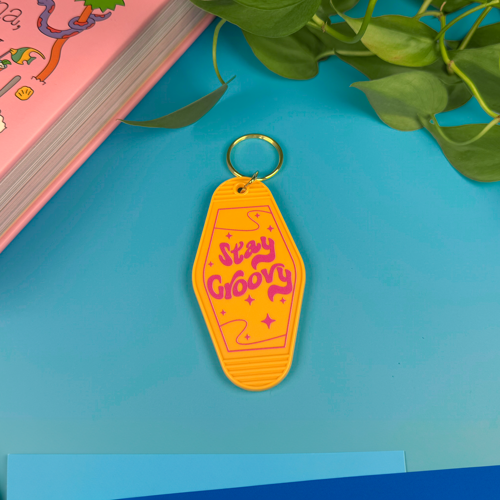 Yellow keychain with 'Stay Groovy' text on a blue surface with a pink book and green leaves.