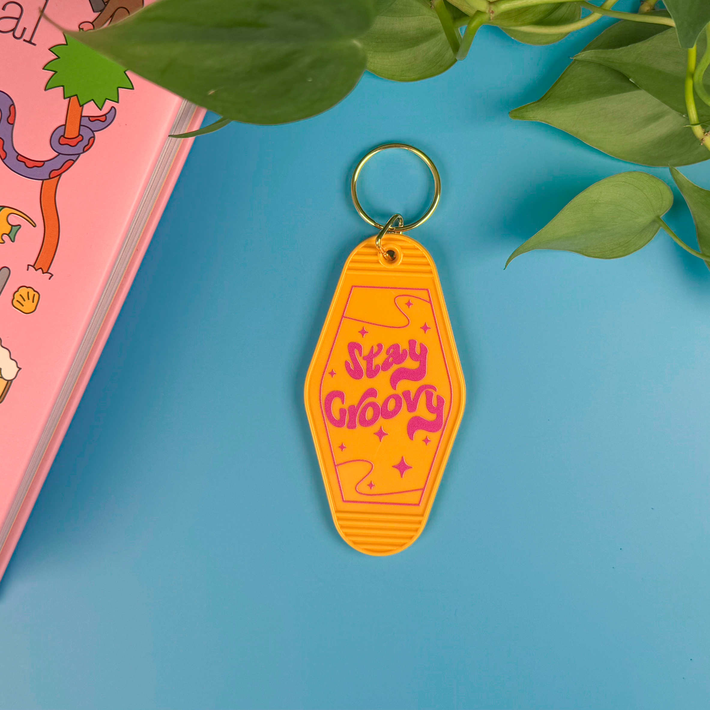 Retro motel-style keychain with 'Stay Groovy' text on a blue background with a pink book and green leaves.